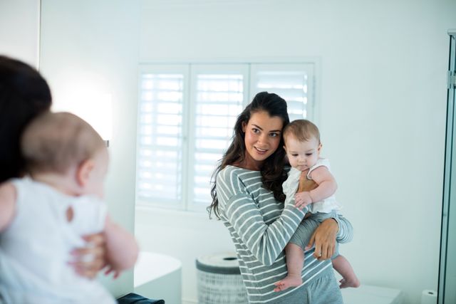 Mother holding baby girl while looking at mirror in home. Ideal for parenting blogs, family lifestyle articles, and advertisements focusing on motherhood and family bonding. Can be used to depict domestic life, parental love, and the joy of raising children.