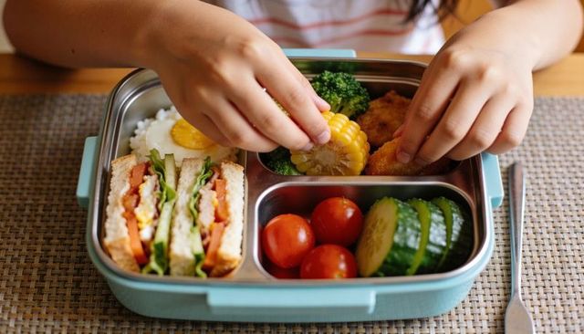 Child's hands reaching bento lunchbox with sandwich, corn, nuggets and fresh veg
