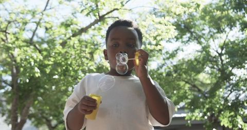 Boy blowing bubbles in sunny backyard holding yellow wand and bottle under trees