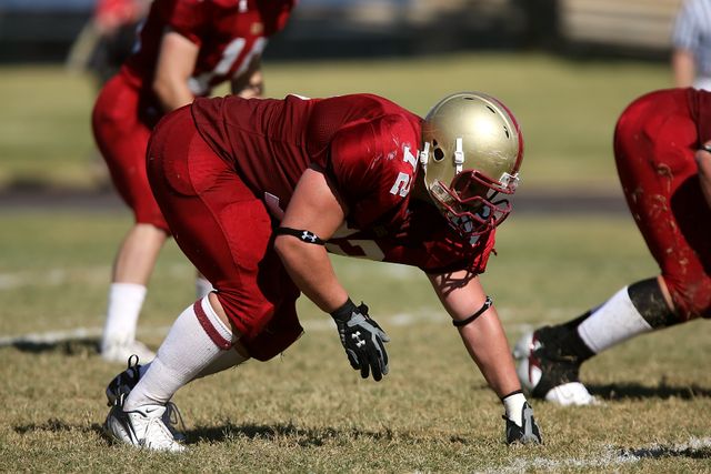 Football player in red uniform and helmet crouching in a defensive stance on a grassy field. Teammates are visible in the background in similar positions. Suitable for sports, team sports, athletic competition, football training, game day promotion, team spirit campaign, and energy.