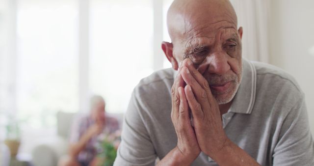 Elderly man sits in living room, looking pensive with his hands on his face. Another senior person is blurred in the background. Useful for themes of aging, loneliness, contemplation, senior care, emotional health, and family relationships.