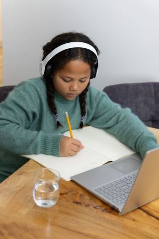 Child studying at home wearing headphones writing in notebook beside laptop