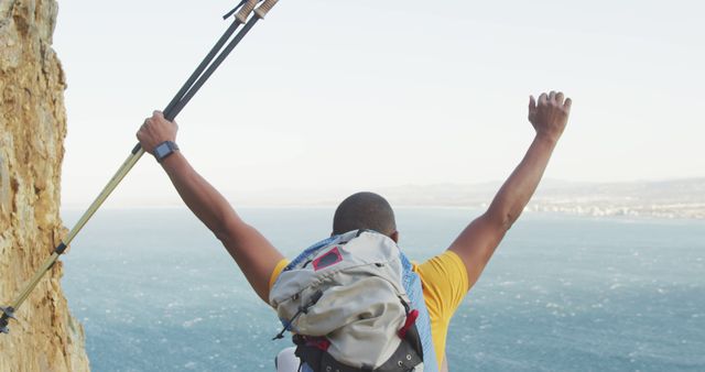 Rear view of biracial man trekking with backpack and walking poles raising arms in victory by sea. Long distance walking, fitness, challenge, nature and healthy outdoor lifestyle.