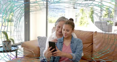 Senior mother embracing daughter while sharing smartphone on sunny living room couch