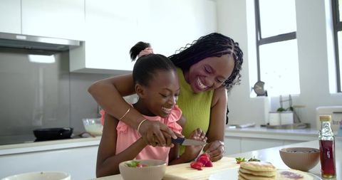 African american mother teaching daughter slicing strawberries smiling in bright kitchen