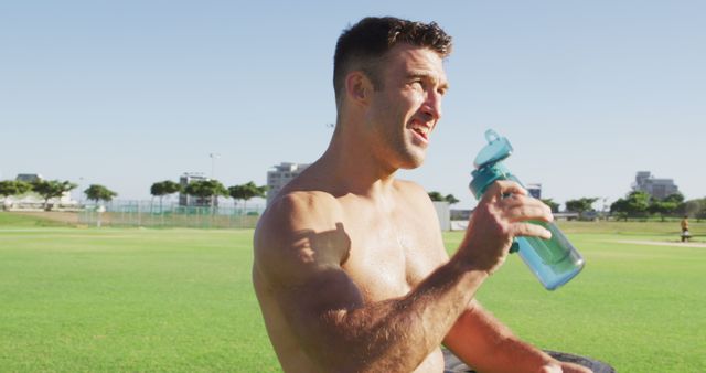 Fit shirtless caucasian man dinking water after exercising outdoors. cross training for fitness at a sports field.