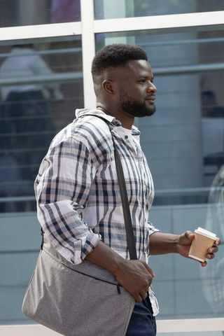 Urban commuter walking with coffee and grey messenger bag in plaid shirt on city street