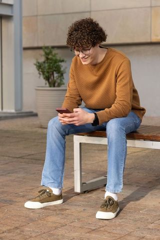 Young man sitting on bench checking smartphone in urban courtyard wearing brown sweater