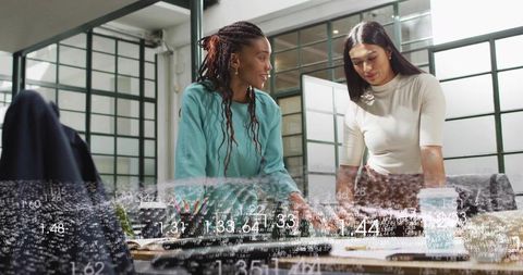Two women collaborating over data visualization and documents on desk in modern glass office