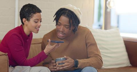 Diverse couple sharing smartphone and coffee on cozy living room couch