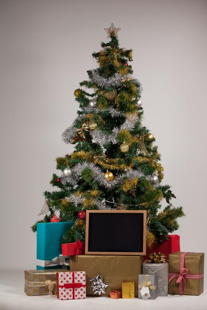 Christmas tree adorned with ornaments and tinsel, surrounded by wrapped gifts and a blank chalkboard. Ideal for holiday promotions, festive greeting cards, and Christmas-themed advertisements.