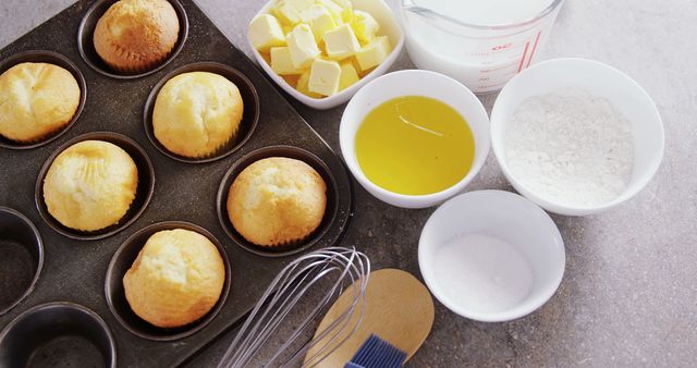 Freshly baked muffins sit in a muffin tin next to ingredients like butter, milk, eggs, flour, and sugar, with copy space. A whisk and a wooden spoon suggest baking preparation or the process of making muffins.