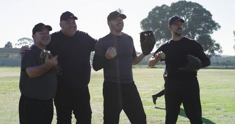 Mature male teammates linking arms and laughing after baseball game on sunlit grass field