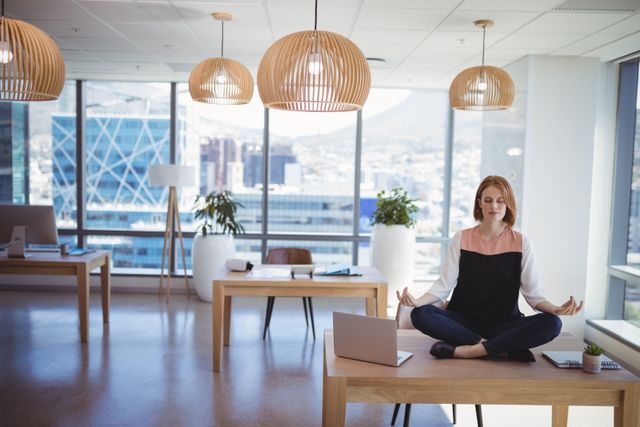 Businesswoman sits cross-legged meditating on an office desk in a modern workspace with panoramic city views. Ideal for articles or marketing materials focusing on workplace wellness, stress relief techniques, work-life balance, or corporate mindfulness programs.