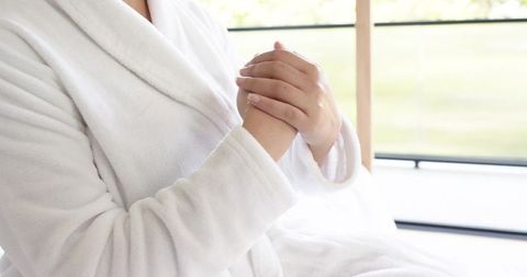 African american woman in plush white bathrobe clasping hands at sunlit balcony window