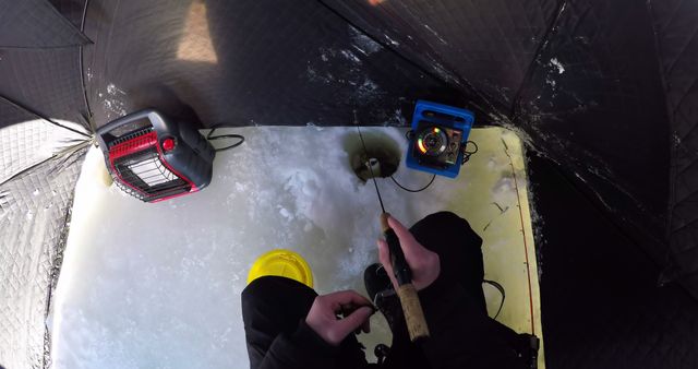 Person ice fishing inside tent, using equipment and looking at ice hole. Perfect for depicting winter outdoor activities, fishing methods, or promoting fishing gear and winter sport equipment.