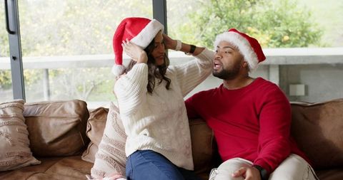 Couple adjusting santa hats on couch sharing cozy holiday moment together