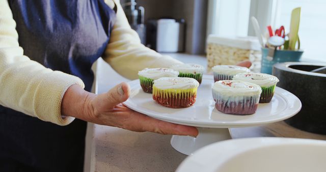 Person holding a tray of cupcakes with frosting in a kitchen. Suitable for content on baking, homemade desserts, kitchen activities, cooking tutorials, or food blogs.