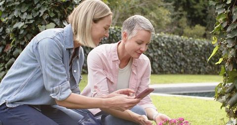 Female friends gardening together while sharing smartphone on backyard lawn near pool