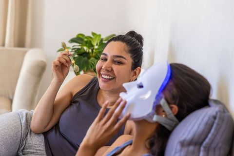 Diverse friends sharing home spa moment using led facial mask and gold face roller