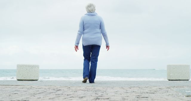 A senior Caucasian woman walks towards the sea on a cloudy day, with copy space. Her solitary stroll along the beachfront evokes a sense of peaceful contemplation and the serenity of nature.