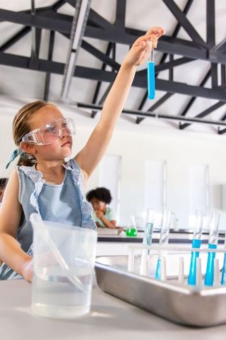 Girl examining blue test tube in classroom science lab wearing goggles and denim dress
