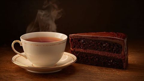 Steaming china teacup on gold-rimmed saucer with glossy chocolate ganache cake slice on warm wood