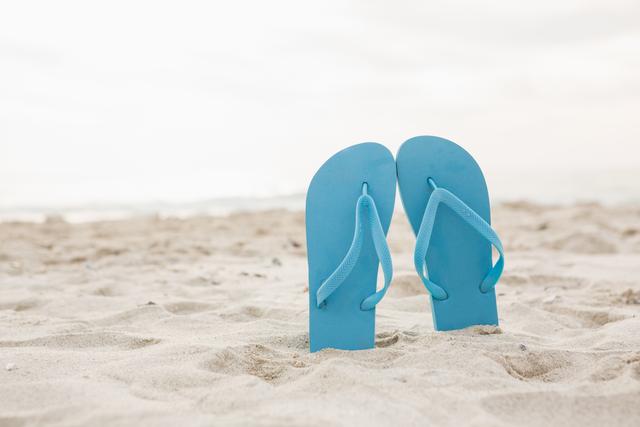 Blue flip-flops standing upright in sand on beach. Evokes feelings of summer and relaxation. Ideal for travel websites, vacation advertisements, beach lifestyle promotions, and summer-themed social media posts.
