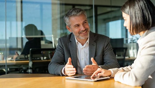 Senior businessman gesturing while advising colleague on tablet at bright office meeting