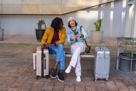 Diverse travelers laughing and checking smartphone while waiting with luggage on bench