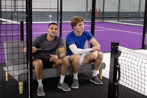 Two men sitting on bench watching indoor paddle court with purple flooring and net