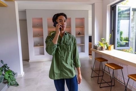 Asian man talking on smartphone in modern minimalist open-plan home with plants