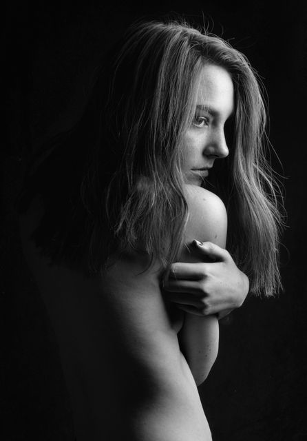 Moody black and white portrait shows a young woman with long hair posing with bare shoulders against a dark background. Her serious expression and thoughtful pose exude a sense of introspection and emotion. Perfect for use in artistic photography collections, emotional storytelling, portrait studies or design themes emphasizing expressive or dramatic moods.