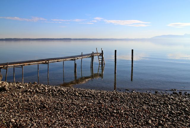 Serene lakeside view showcasing a wooden pier extending into calm water, surrounded by a rocky shore and clear blue sky. Perfect for use in projects related to relaxation, nature retreats, travel destinations, and promoting mindfulness. Ideal background for websites, brochures, and advertisements focusing on peaceful getaways.
