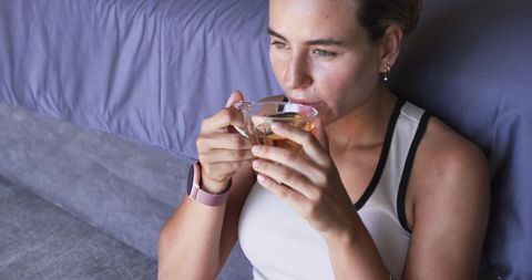 Young woman sipping herbal tea on bed wearing rose-gold smartwatch and casual tank top