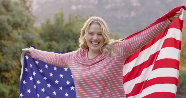 Smiling woman holding American flag with outstretched arms surrounded by greenery and mountains. Ideal for themes on patriotism, Independence Day celebrations, American identity, freedom, or outdoor activities.