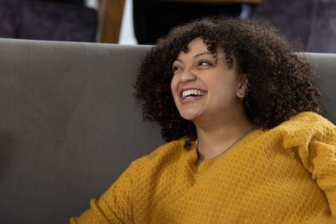 Laughing african american woman on sofa wearing mustard sweater, cozy relaxed mood