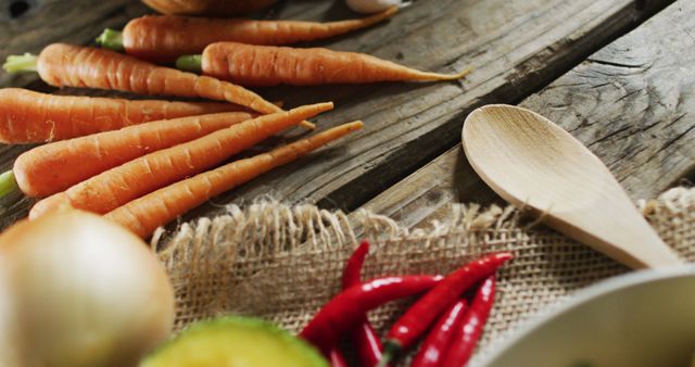 Fresh vegetables including carrots, onion, and chili peppers are placed on a rustic wooden table next to a wooden spoon. Perfect for use in content related to healthy eating, organic cooking, farm-to-table concepts, and culinary blogs.
