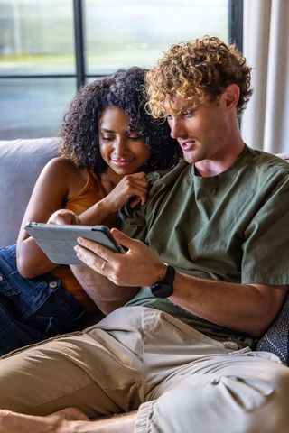 Sunlit diverse couple relaxing on sofa watching tablet together