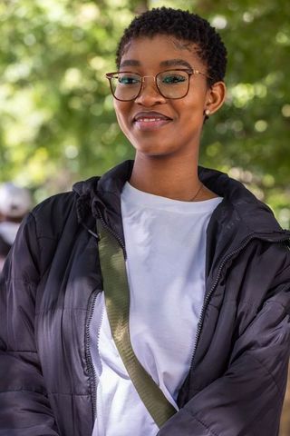 African woman smiling in sunlit park wearing eyeglasses and black puffer jacket