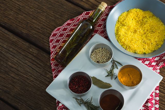 This image shows a close-up of a bowl of yellow rice accompanied by various spices and a bottle of oil on a wooden table. Ideal for use in culinary blogs, recipe websites, cooking tutorials, and food-related advertisements. The vibrant colors and rustic setting make it perfect for promoting gourmet and home-cooked meals.