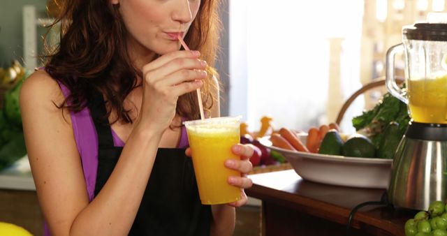 Woman enjoying fresh orange juice in a kitchen surrounded by fruits and vegetables. Ideal for concepts related to healthy living, kitchen appliances, fresh drinks, and nutrition. Useful for health blogs, juicing recipes, and promotional material for kitchenware.