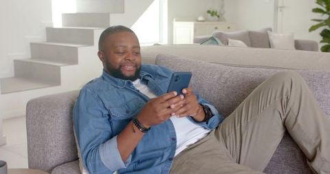 African american man reclining on sofa using smartphone in modern bright living room
