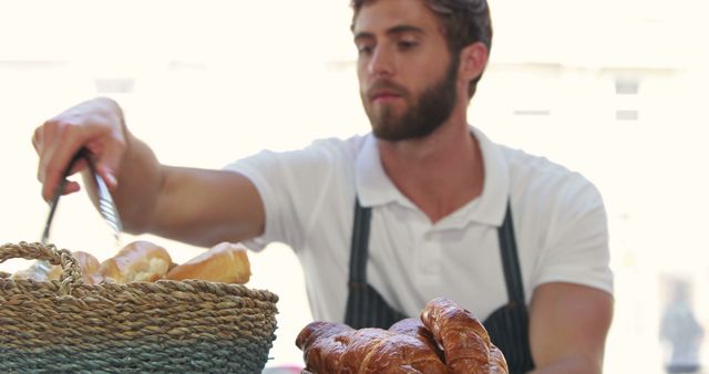 Young male baker is arranging fresh pastries, such as baguettes and croissants, in bakery. He is in a white shirt with an apron, displaying bread in woven baskets. Perfect for illustrating topics on small businesses, artisanal baking, or food industry professions.