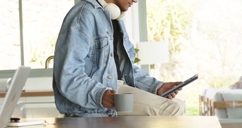 Mid-adult man sitting on kitchen counter using tablet and holding mug, denim jacket