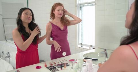 Diverse women styling hair and applying makeup at bathroom vanity in evening dresses