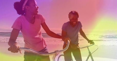 Couple walking bikes along sunlit beach laughing, enjoying romantic coastal ride