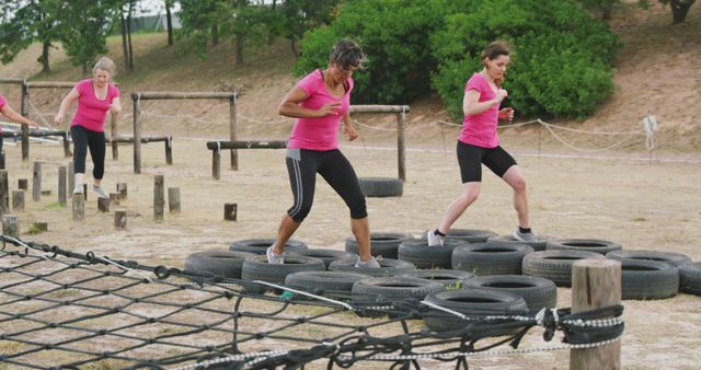 Determined caucasian female friends in pink t shirts crossing logs and tyres at bootcamp training. Female fitness, friendship, challenge and healthy lifestyle.