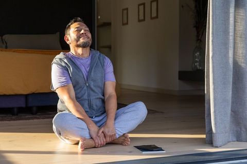 Middle-aged asian man relaxing barefoot on sunlit floor in minimalist bedroom with tablet