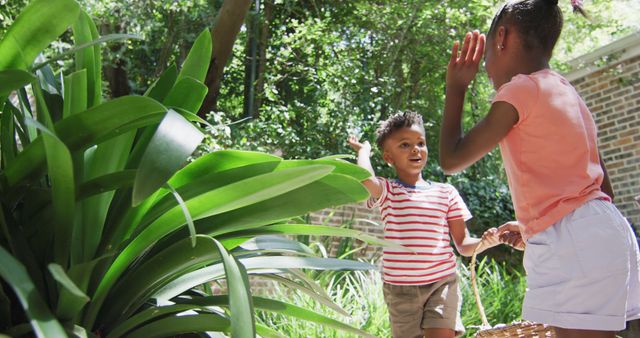 Children exploring garden with basket, interacting next to plants in sunlight. Useful for topics related to childhood, outdoor activities, family bonding times, nature exploration, or summer activities.
